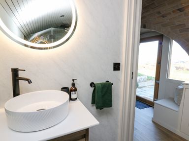A bathroom sink with a round mirror and dark faucet next to an open door showing a small hallway with a window seat at Tan y Wawr Onnen in Llanfair Caereinion
