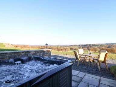 An outdoor hot tub with bubbling water and a wooden table with four chairs on gravel overlooking a rural landscape at Tan y Wawr- Onnen in Llanfair Caereinion