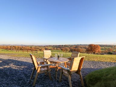 An outdoor patio area with wooden chairs and a round table overlooking a field with trees at Tan y Wawr- Onnen in Llanfair Caereinion