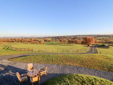 An outdoor sitting area with wooden chairs and a table on gravel facing fenced green fields and trees at Tan y Wawr- Onnen in Llanfair Caereinion