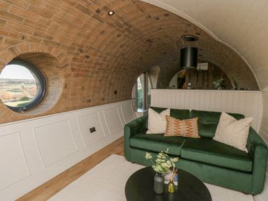 A living area with a green sofa and cushions a round window and a round black table with decorative items at Tan y Wawr- Onnen in Llanfair Caereinion