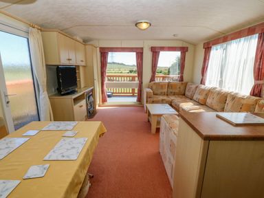 A living area with an L-shaped sofa a coffee table a dining table with placemats a television unit and large windows at Gorphwysfa Caravan in Tregele near Cemaes Bay