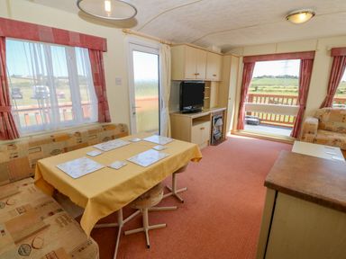 A living area with a dining table, bench seating, television on a cabinet, and windows with red curtains at Gorphwysfa Caravan Tregele near Cemaes Bay