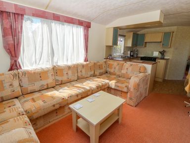 A living room with a patterned corner sofa and coffee table next to a small kitchen at Gorphwysfa Caravan in Tregele near Cemaes Bay