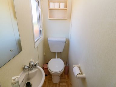A small bathroom with a toilet a sink a mirror a window and shelves with toilet paper at Gorphwysfa Caravan in Tregele near Cemaes Bay