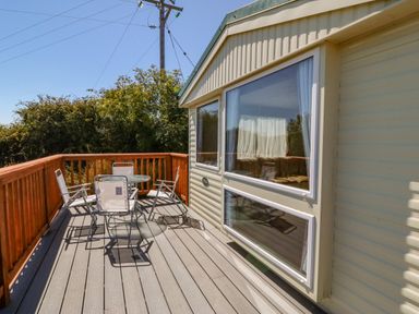 A wooden deck with a round glass table and four metal chairs outside a caravan at Gorphwysfa Caravan in Tregele near Cemaes Bay
