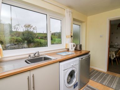 A kitchen with a sink, washing machine, small fridge, kettle, toaster, and window showing a garden at Llwynon in Goginan near Aberystwyth