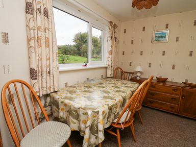 A dining room with wooden chairs a table with a patterned tablecloth a sideboard and a window showing greenery outside at Llwynon in Goginan near Aberystwyth