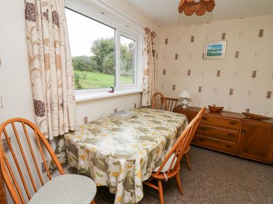 A dining room with wooden chairs a table with a patterned tablecloth a sideboard lamp and a window showing a garden at Llwynon in Goginan near Aberystwyth