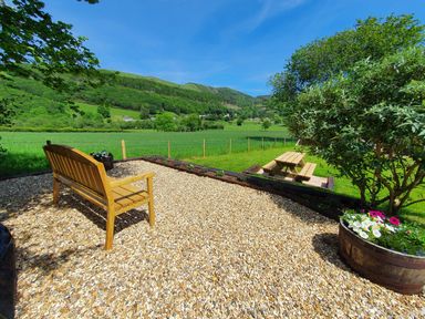 An outdoor seating area with a wooden bench a flower pot a picnic table and a tree overlooking green fields and hills at Llwynon in Goginan near Aberystwyth