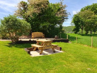 A wooden picnic table on a grass lawn with a wooden bench and trees in the background at Llwynon in Goginan near Aberystwyth