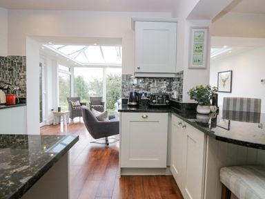 A kitchen with black countertops and white cabinets opening to a seating area with chairs and a glass conservatory at Holme Fell in Coniston