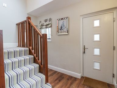 A hallway with a striped carpeted staircase wooden railing framed artwork window with a floral blind white door and a brown doormat at Holme Fell in Coniston
