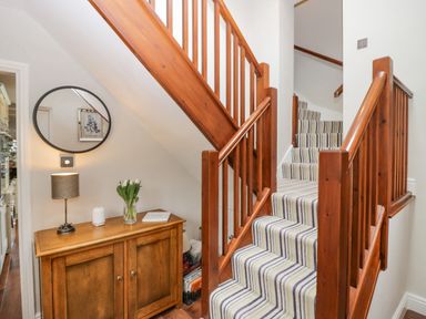 A staircase with striped carpet and wooden railing next to a wooden cabinet with a lamp and flowers at Holme Fell in Coniston