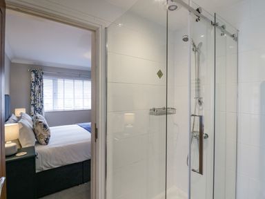 A bathroom with a glass shower enclosure and a bedroom with a bed and nightstand visible through the door at Holme Fell in Coniston