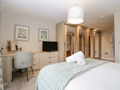 A bedroom with a bed, rolled towels, a TV on a dresser, a chair at a dressing table, framed pictures, and wardrobes with mirrored doors at Holme Fell in Coniston