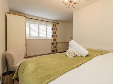 A bedroom with a bed covered with a green blanket and towels on top a beige chair a cream wardrobe cream dresser and window with white blinds at Holme Fell in Coniston