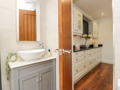 A small bathroom with a white oval sink on a gray cabinet and a mirror above it next to a hallway with white cabinets and wooden floor at Holme Fell in Coniston