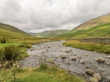 A river with rocks flowing through a valley between grassy hills with a stone wall on one side at Holme Fell in Coniston