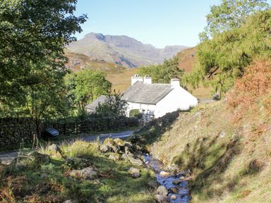 A cottage by a stream with trees and hills in the background at Holme Fell in Coniston