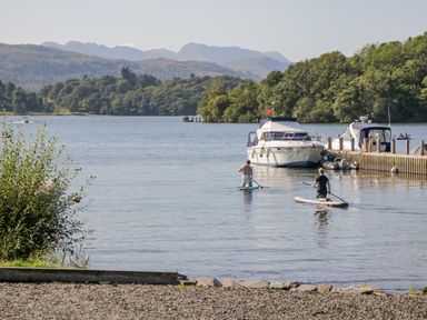 A lake with two people paddleboarding near boats and a dock surrounded by trees and hills at Holme Fell in Coniston