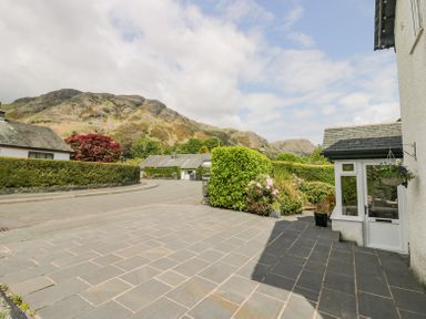 A paved driveway with bushes and flowers next to a white house with mountains in the background at Holme Fell in Coniston