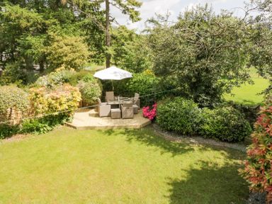 An outdoor patio with table chairs and umbrella surrounded by trees and shrubs at Holme Fell Coniston