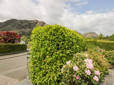 A garden with green shrubs and pink flowers near a road with hills in the background at Holme Fell Coniston