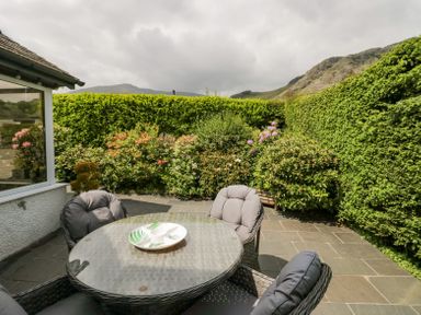 A garden patio with a round glass table and cushioned chairs surrounded by hedges and flowering plants at Holme Fell in Coniston