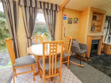 A dining area with a round wooden table and four chairs next to a window with striped curtains and a gray rocking chair beside a fireplace at Rosehaven in Tegryn near Crymych