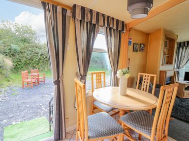 A dining area with a round wooden table and four chairs next to windows with curtains and an open door showing two wooden chairs outside at Rosehaven in Tegryn near Crymych