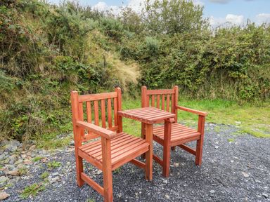 Two wooden chairs with a small wooden table between them on a gravel area surrounded by green grass and bushes at Rosehaven in Tegryn near Crymych