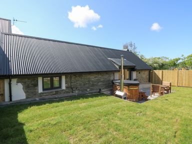 An outdoor area with grass a stone building with a corrugated roof a wooden hot tub and wooden patio furniture at The Parlour in Llangoedmor near Cardigan