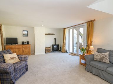 A living room with two armchairs a wooden cabinet a wood stove and glass doors with yellow curtains at Cattle Tree Cottage in Llangoedmor near Cardigan
