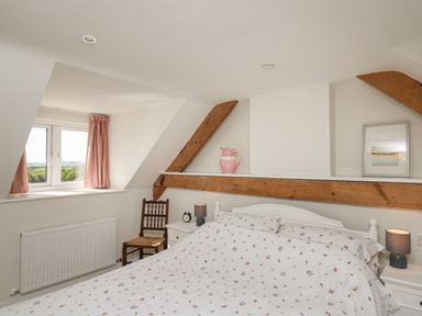 A bedroom with a bed with floral bedding two bedside tables with lamps a chair under a window with pink curtains and wooden beams on walls at Cattle Tree Cottage in Llangoedmor near Cardigan