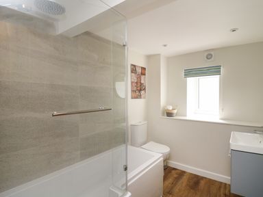 A bathroom with a bathtub shower, toilet, window with striped blind, sink, and wooden floor at Cattle Tree Cottage in Llangoedmor near Cardigan