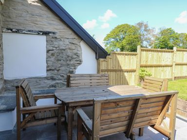An outdoor patio with a wooden table and four wooden chairs surrounded by a stone wall and wooden fence at Cattle Tree Cottage in Llangoedmor near Cardigan