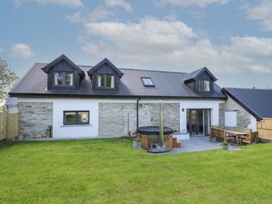 An exterior view of a stone and white house with a black metal roof and three dormer windows a grassy yard a circular hot tub and wooden outdoor furniture at Cattle Tree Cottage in Llangoedmor near Cardigan