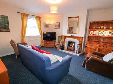 A living room with a blue sofa, brown armchairs, a wooden cabinet with plates and cups, a fireplace with a mirror above it, a television on a wooden stand, and yellow curtains on the window at 17 Buninver Road in Gortin