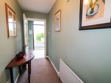 A narrow hallway with green walls a wooden table with a bowl on it paintings on the walls and an open door leading outside at 17 Buninver Road Gortin