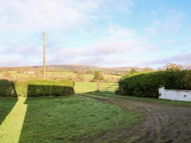 A rural driveway with grass and hedges leading to a metal gate with fields and hills in the background at 17 Buninver Road Gortin