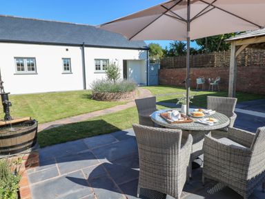 A garden patio with woven chairs around a table under an umbrella with a white building and a grassy area in the background at Catamouse in Milford Haven