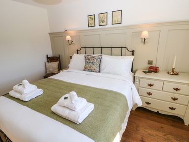 A bedroom with a bed with white linens and a green blanket two stacks of towels on the bed a wooden chair with a cushion a cream dresser with books and a candle and framed pictures on the wall at Catamouse in Milford Haven