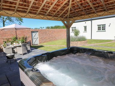 An outdoor hot tub under a wooden gazebo with string lights a patio dining set with umbrella a brick wall and a lawn at Catamouse in Milford Haven