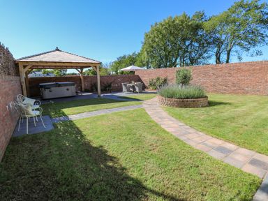 A garden with grass a brick circular flower bed a paved pathway two chairs under a wooden gazebo and a dining table with umbrella at Catamouse in Milford Haven