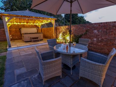 An outdoor patio with a round table and four chairs under an umbrella and a hot tub with string lights under a wooden gazebo at Catamouse in Milford Haven
