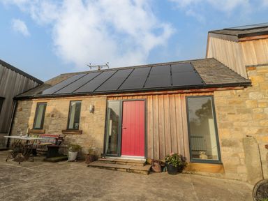 A stone and wood cottage exterior with solar panels on the roof a red door and patio furniture at High Barns Cottage in Longhorsley near Morpeth