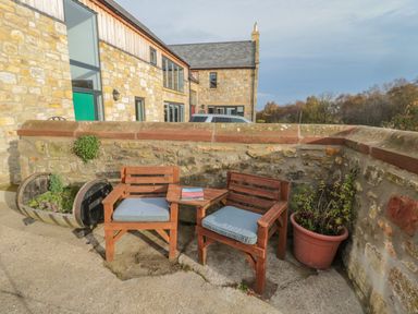 A small outdoor seating area with two wooden chairs with blue cushions and a triangular wooden table between them at High Barns Cottage in Longhorsley near Morpeth
