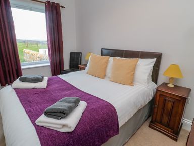 A bedroom with a double bed two beige pillows folded towels on a purple runner window with maroon curtains a wooden nightstand with a yellow lamp and a black chair at High Barns Cottage in Longhorsley near Morpeth