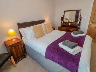 A bedroom with a bed with white linens and purple runner with towels two wooden bedside tables with lamps a wooden dresser with mirror at High Barns Cottage in Longhorsley near Morpeth